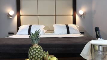 Ett hotellrum med en säng, två kuddar, en sänggavel, sänglampor och en fruktskål med ananas, kiwi, apelsin och melon på nattduksbordet.