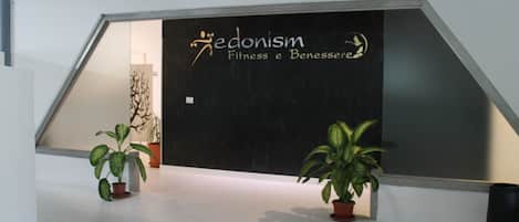 Un interior moderno con una pared negra que muestra el logotipo y el nombre 'Pedonism Fitness e Benessere'. Hay dos plantas en macetas sobre el suelo.