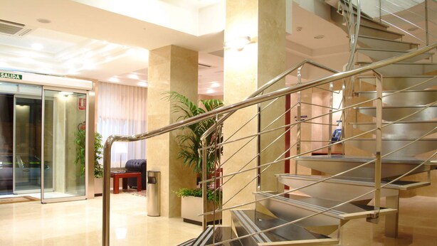 Um hall de hotel moderno com entrada de vidro, escada com guarda-corpo de metal e balcão de recepção.