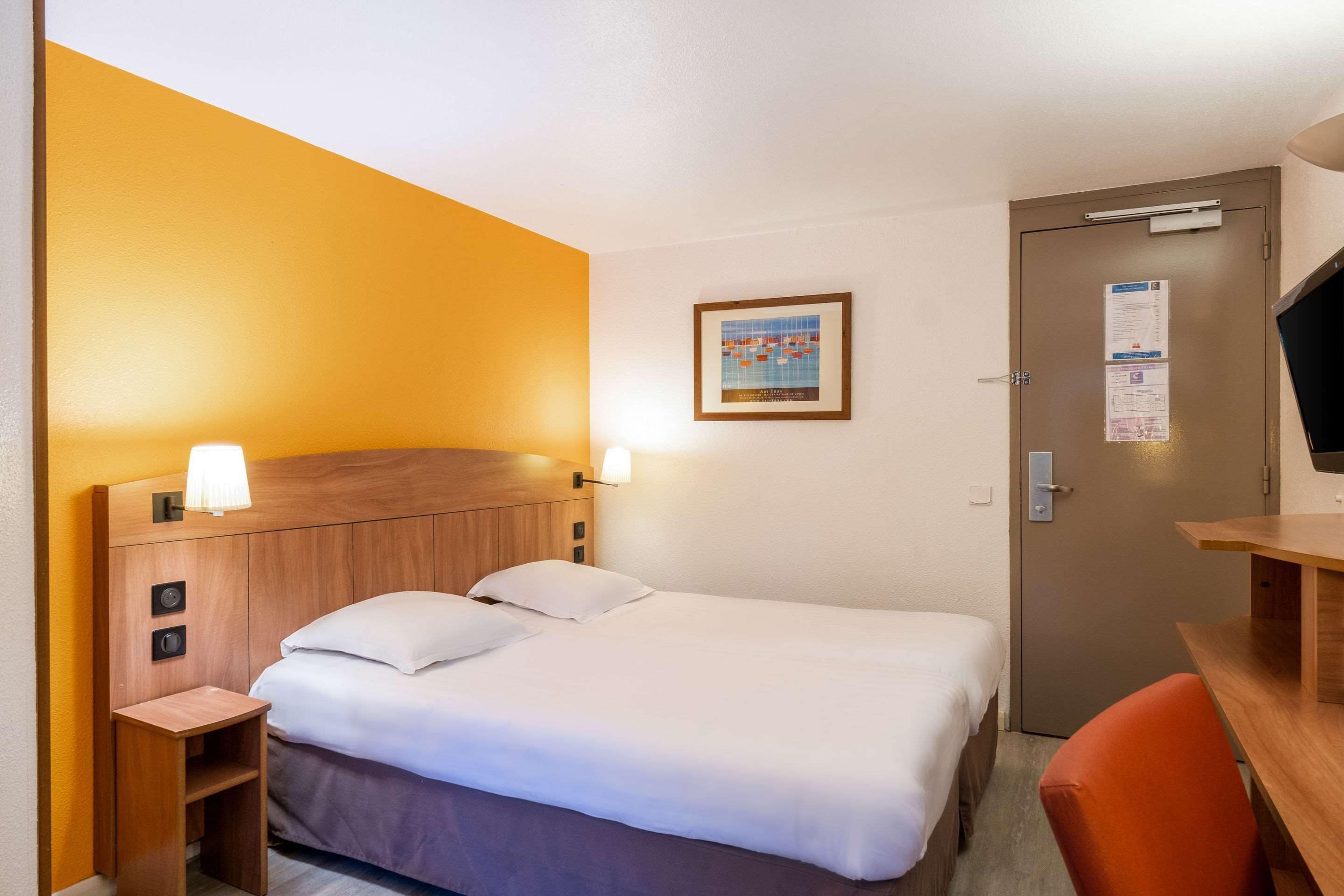 Una habitación de hotel con cama, mesita de noche, lámpara y una puerta con un tablón de anuncios.