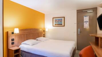 Una habitación de hotel con cama, mesita de noche, lámpara y una puerta con un tablón de anuncios.