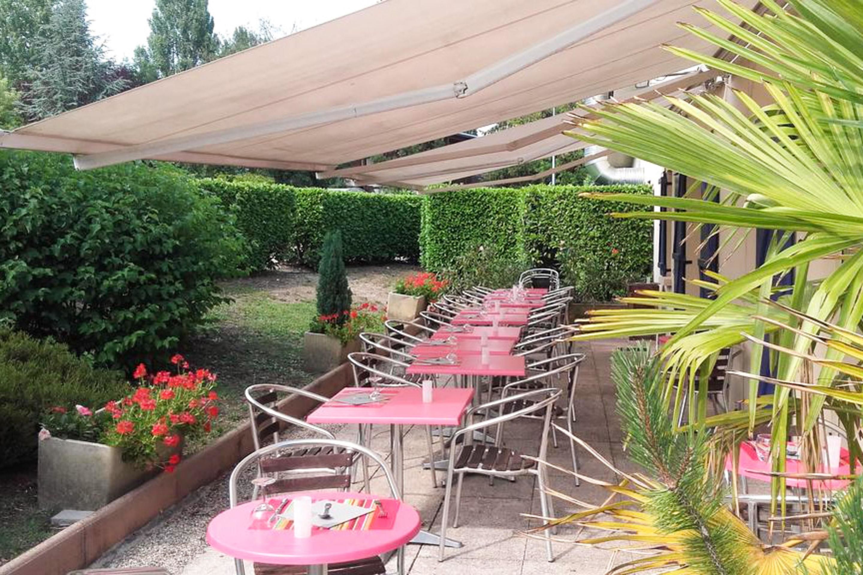 An outdoor patio with metal chairs and tables, surrounded by greenery and a hedge.