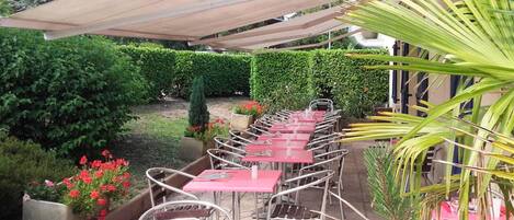 An outdoor patio with metal chairs and tables, surrounded by greenery and a hedge.