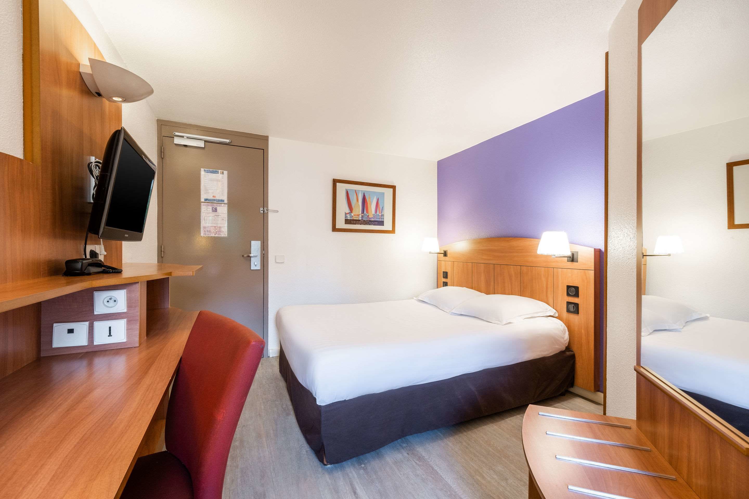 A hotel room with a bed, a desk with a chair, a TV, and a mirror.