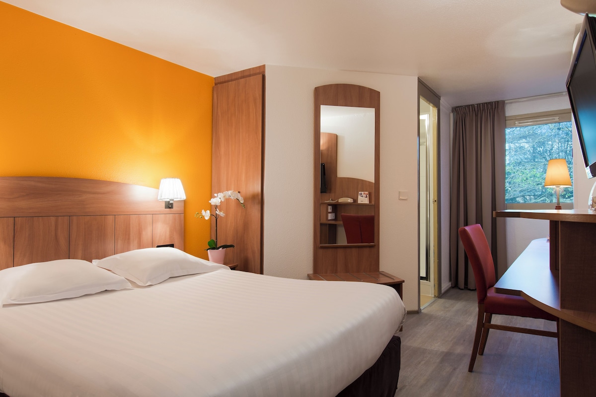 A hotel room with a large bed, a desk, a chair, a TV, and a window with curtains.