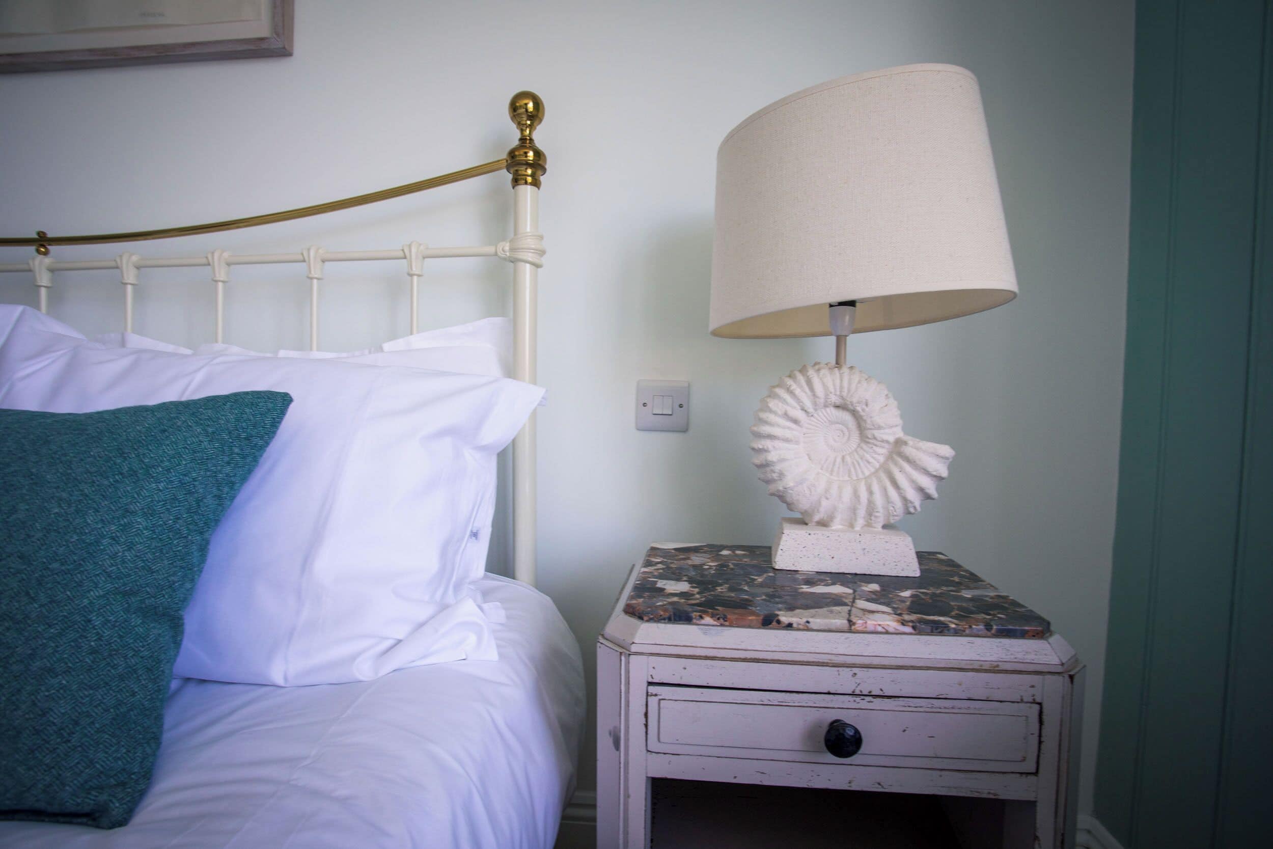 A bed with white bedding and a teal pillow, a white nightstand with a lamp and a decorative shell, and a wall switch.