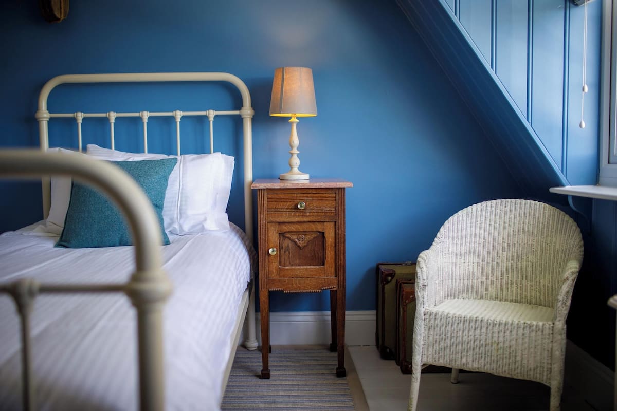 A bedroom with a bed, a nightstand with a lamp, a wicker chair, and a suitcase.