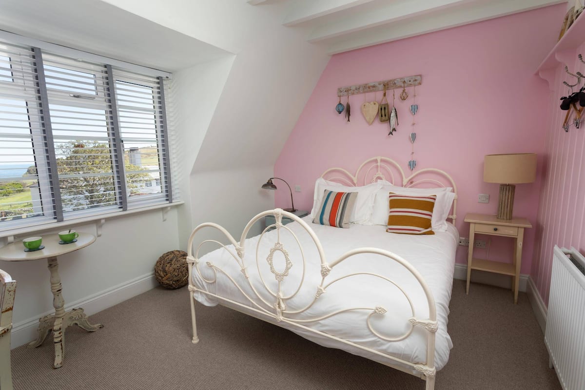 A bedroom with a white metal bed, pink walls, a window with blinds, a small table with a lamp, and a radiator.