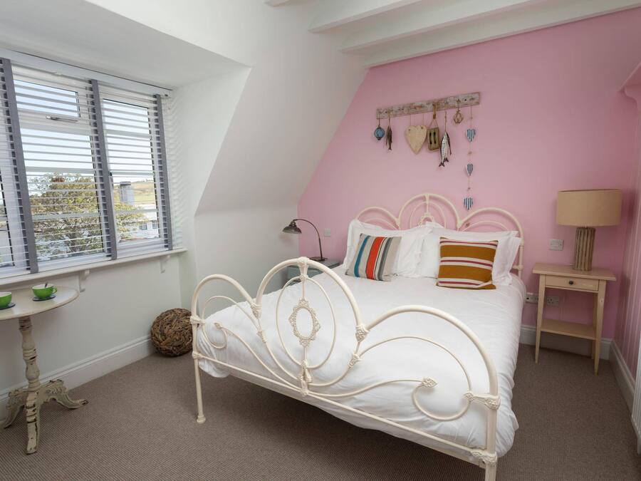 Ein Schlafzimmer mit einem weißen Metallbett, rosafarbenen Wänden, einem Fenster mit Jalousien, einem kleinen Tisch mit Lampe und einer Heizung.