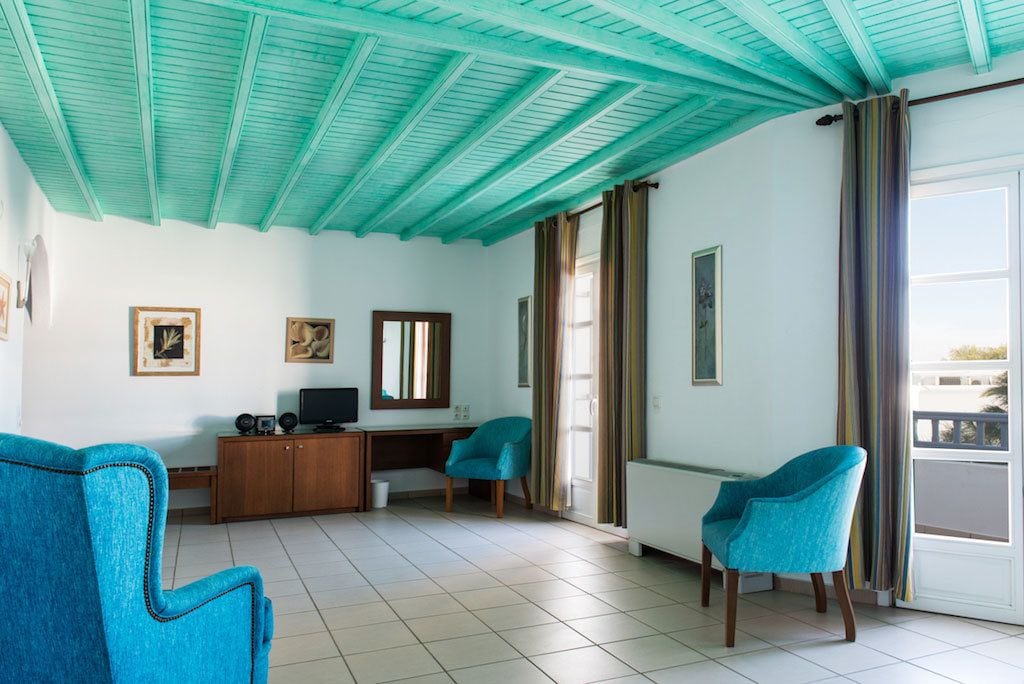 A living room with teal ceiling beams, a wooden desk, two blue chairs, and a large window with curtains.