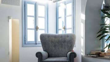 Ein Zimmer mit grauem Sessel, zwei Fenstern mit blauen Rollläden, einer hohen Stehlampe und einer Blumentopf-Pflanze.