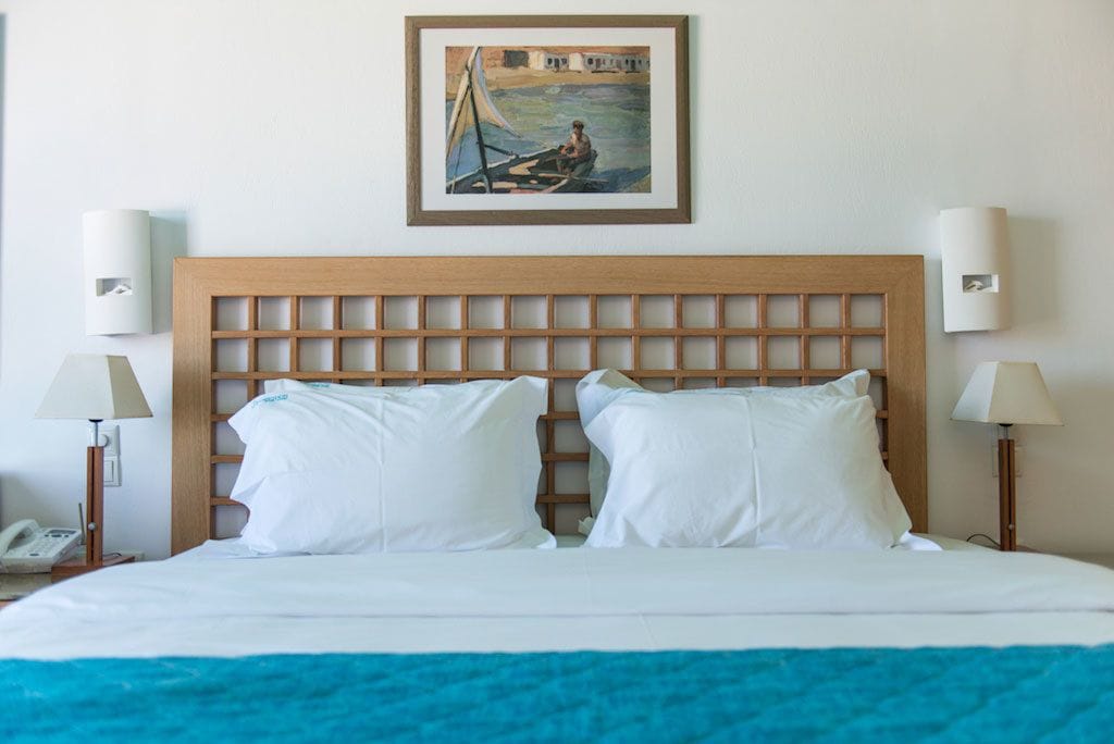 A hotel room with a wooden headboard, white bedding, two bedside lamps, and a framed painting above the bed.