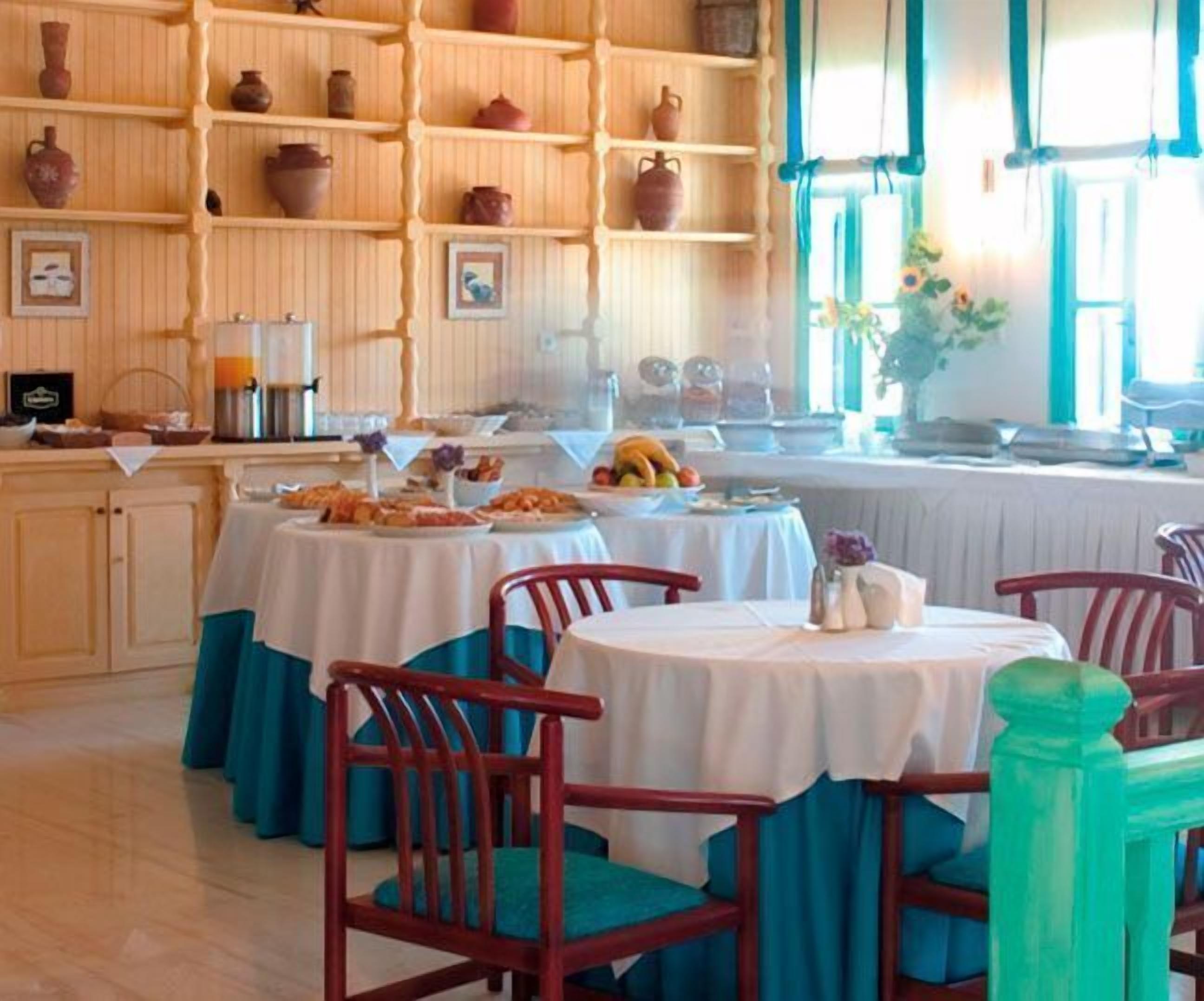 A dining area with round tables covered in white tablecloths, surrounded by red chairs. There are shelves with pottery and framed pictures on the wall.
