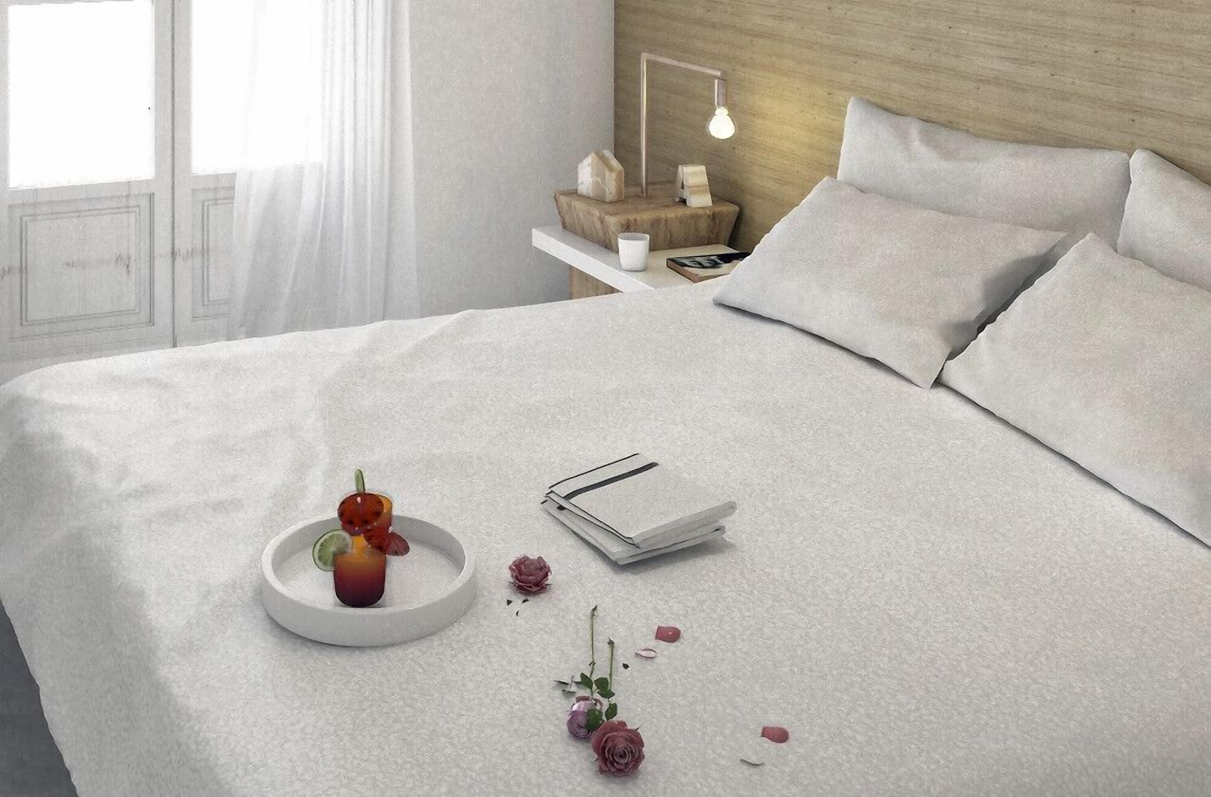 Una cama bien hecha con almohadas, una mesita de noche con lámpara y una bandeja con una bebida y pétalos de rosa.