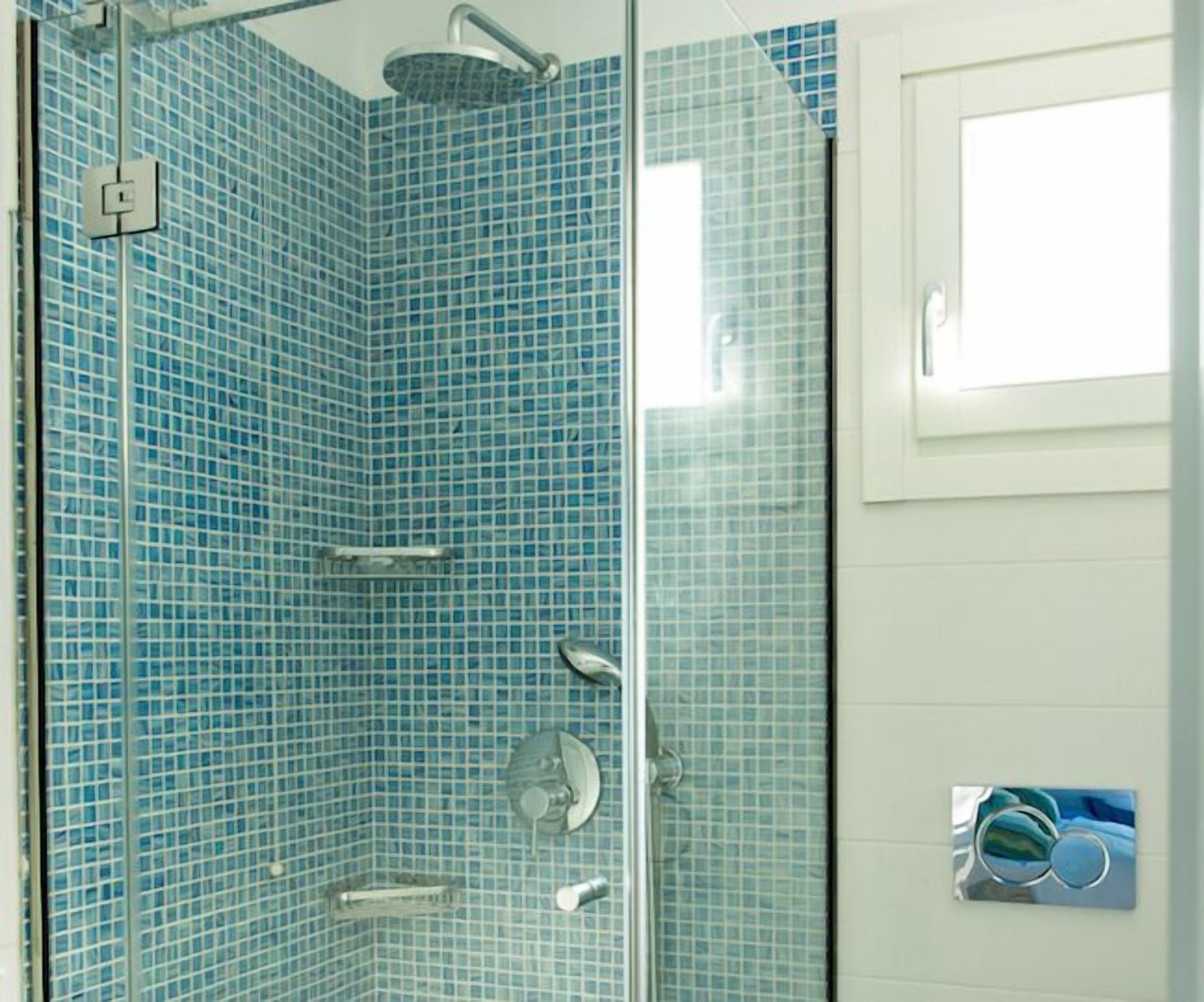 Una ducha con cerramiento de vidrio, azulejos de mosaico azules, una puerta de vidrio y un ventanal pequeño.