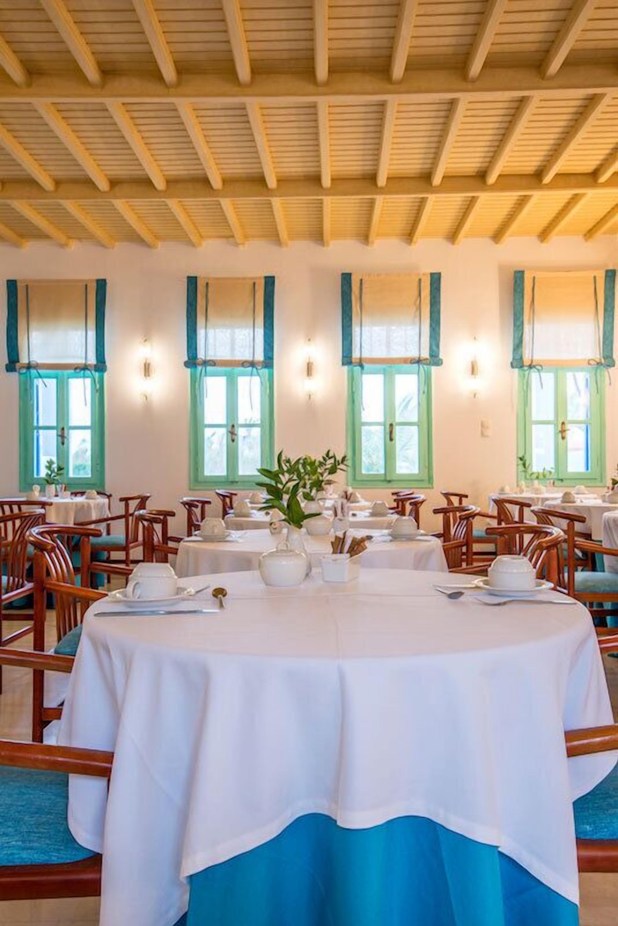 A dining area with round tables set for service, wooden chairs, and a ceiling with wooden beams.