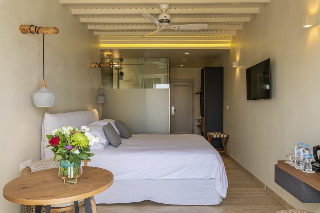 A hotel room with a bed, a round wooden table with a vase of flowers, a ceiling fan, and a TV mounted on the wall.
