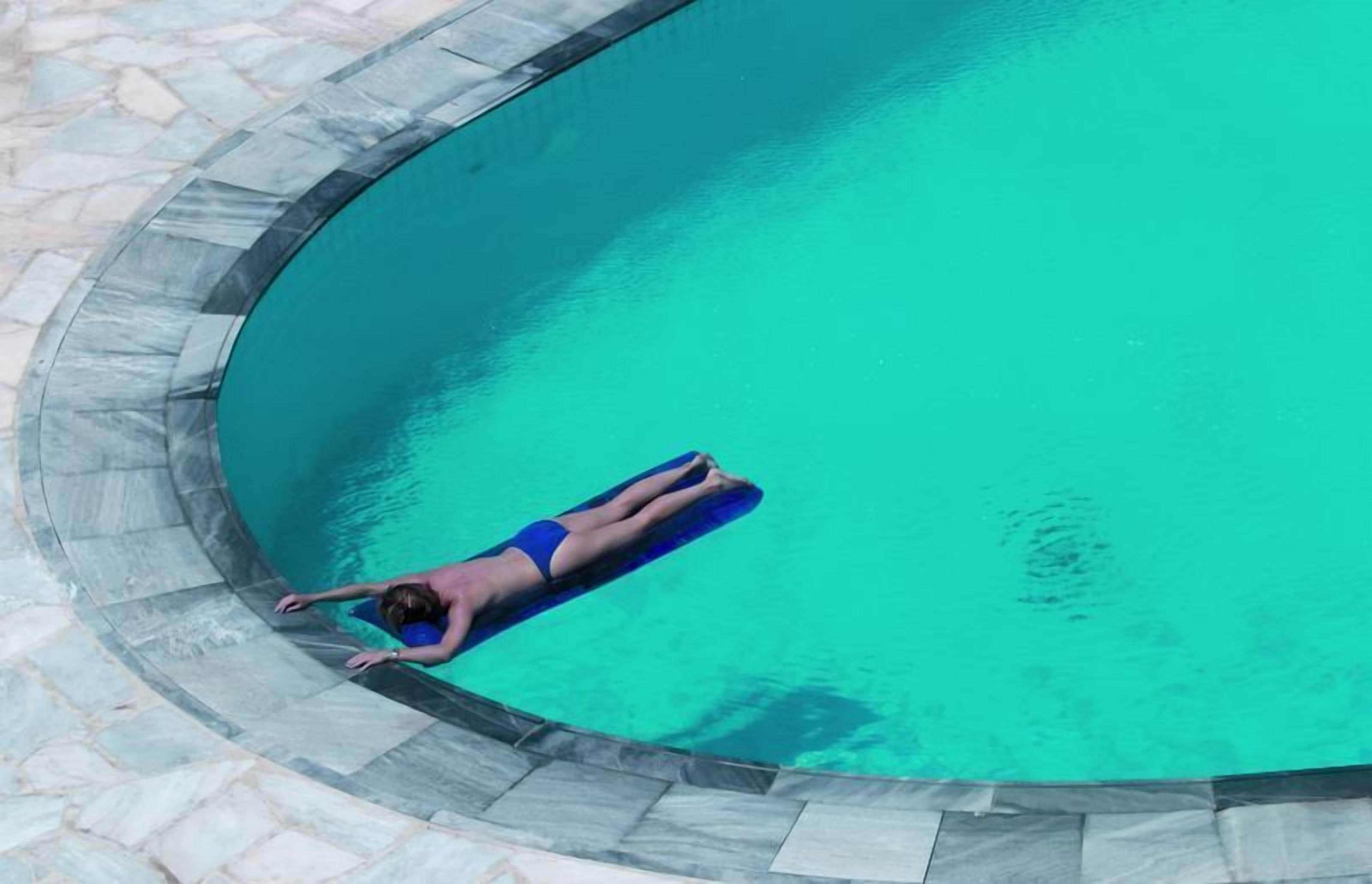 A person floating on a blue floatation device in a clear blue pool with a stone border.