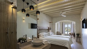 A modern bedroom with a large bed, a built-in vanity with a bowl sink, and pendant lighting. - Poseidon Hotel & Suites (Mykonos)