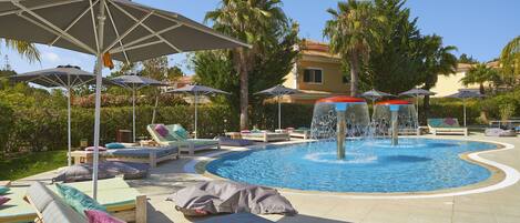 Un área de piscina con sillones de descanso, sombrillas y una fuente de agua.