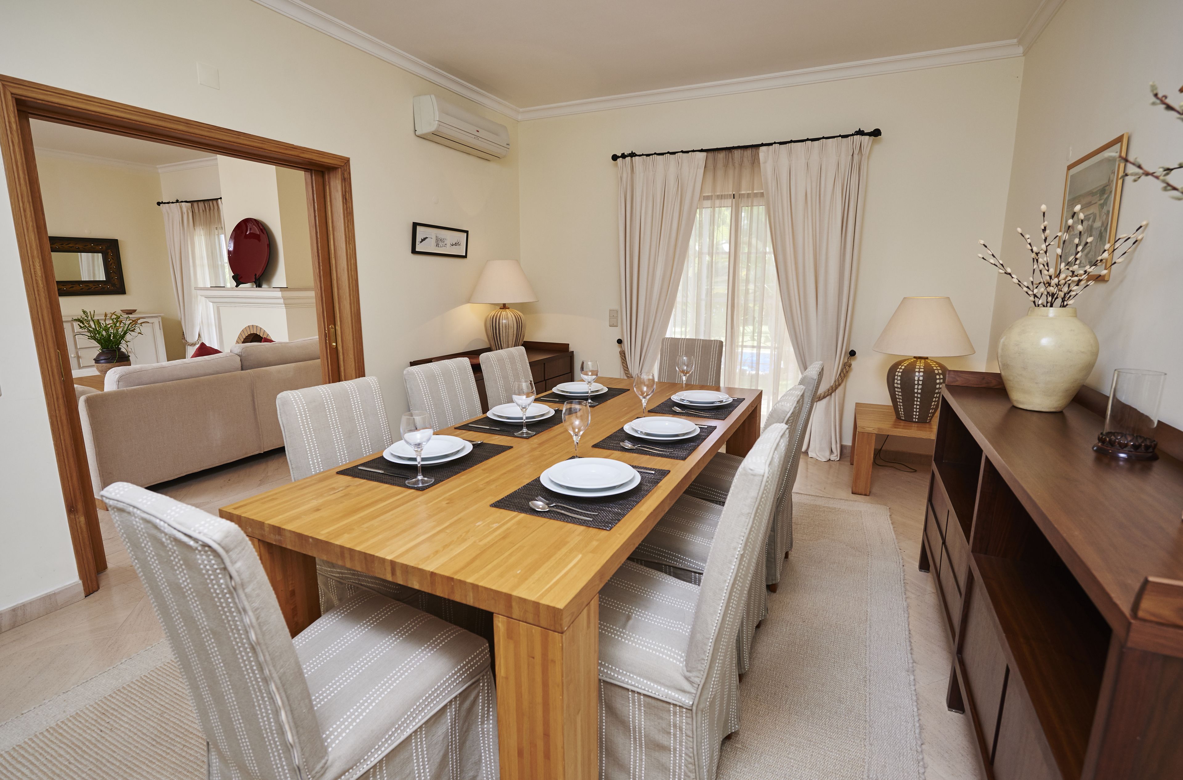 A dining area with a wooden table set for six, surrounded by upholstered chairs. A sofa and a small table with a lamp are visible in the background.