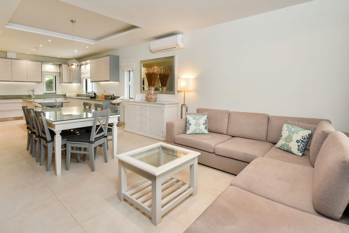 A modern living room with a beige sofa, a glass coffee table, and a dining area with a glass table and chairs.