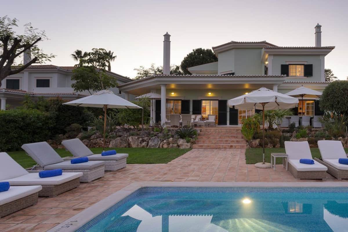 A poolside area with lounge chairs and umbrellas, a stone pathway leading to a house, and a clear blue pool.
