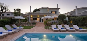 Un espace aménagé au bord de la piscine, avec des chaises longues et des parasols, un chemin de pierre menant à une maison, et une piscine à l’eau d’un bleu limpide.