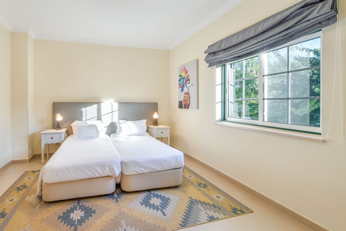 A double bed with two bedside tables, a window with a view of greenery, and a patterned rug.