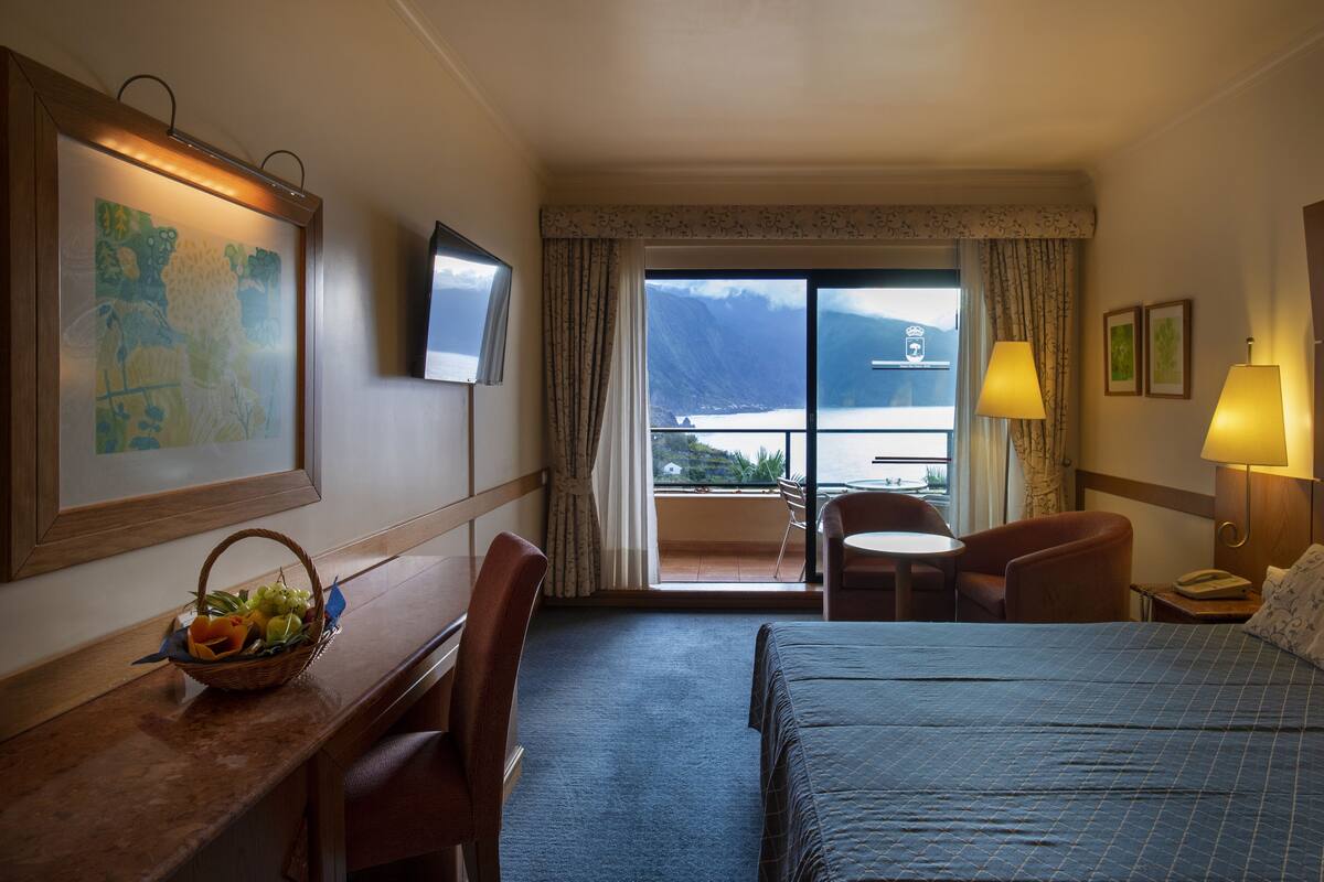Une chambre d’hôtel comprenant un lit, un bureau avec un panier de fruits, une chaise, une table, une lampe et une vue sur la mer et les montagnes, visible à travers une grande fenêtre.