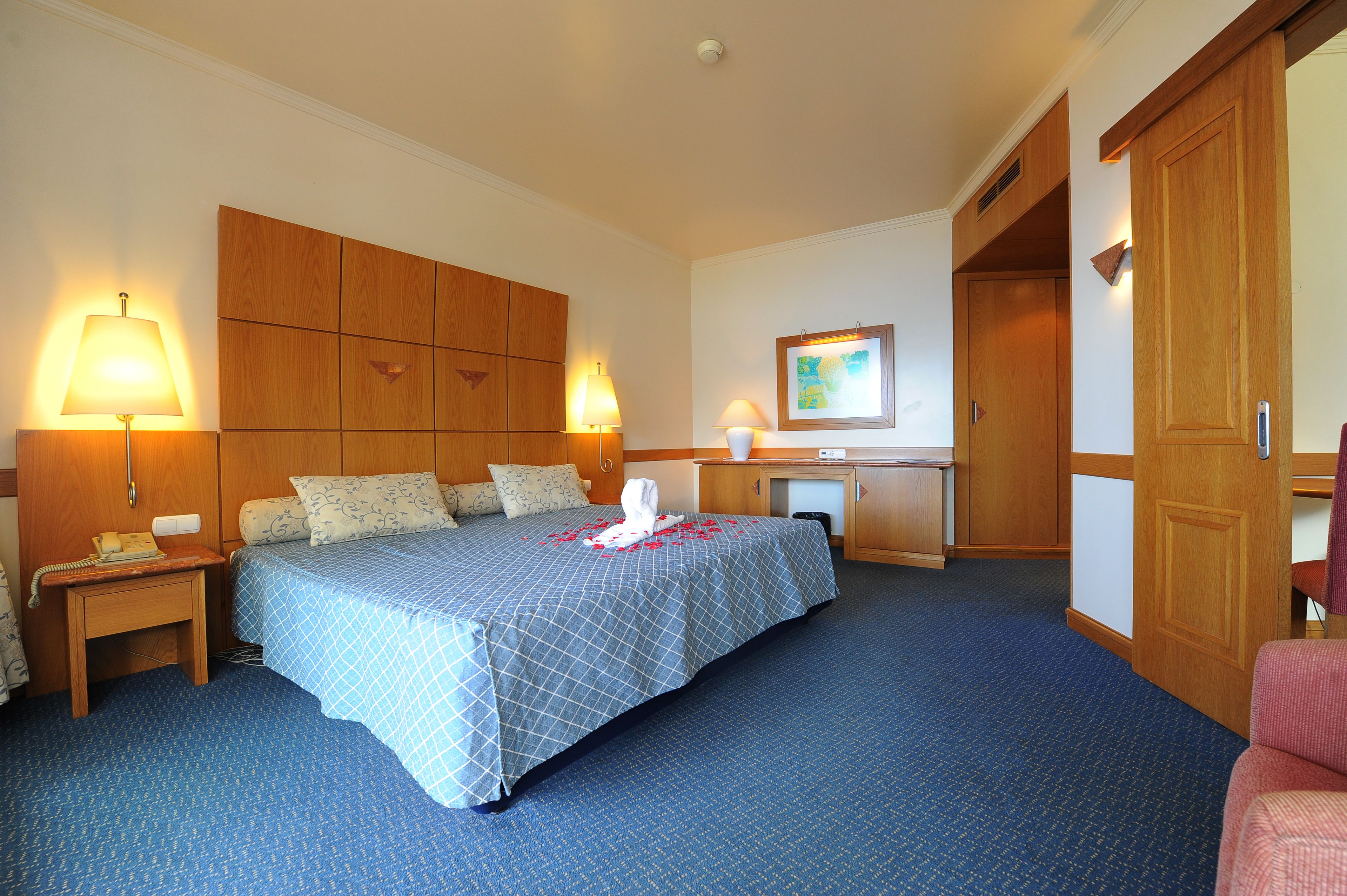 Hotelkamer met een groot bed, twee bedlampen, een houten garderobe, een bureau met stoel, en een schilderij aan de muur.