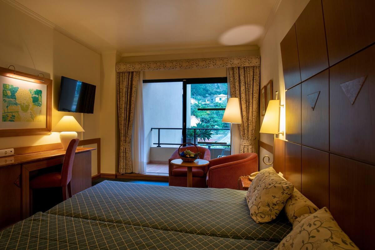 Une chambre d’hôtel comprenant un lit, un bureau, une chaise, une petite table avec un panier, et un téléviseur fixé au mur.