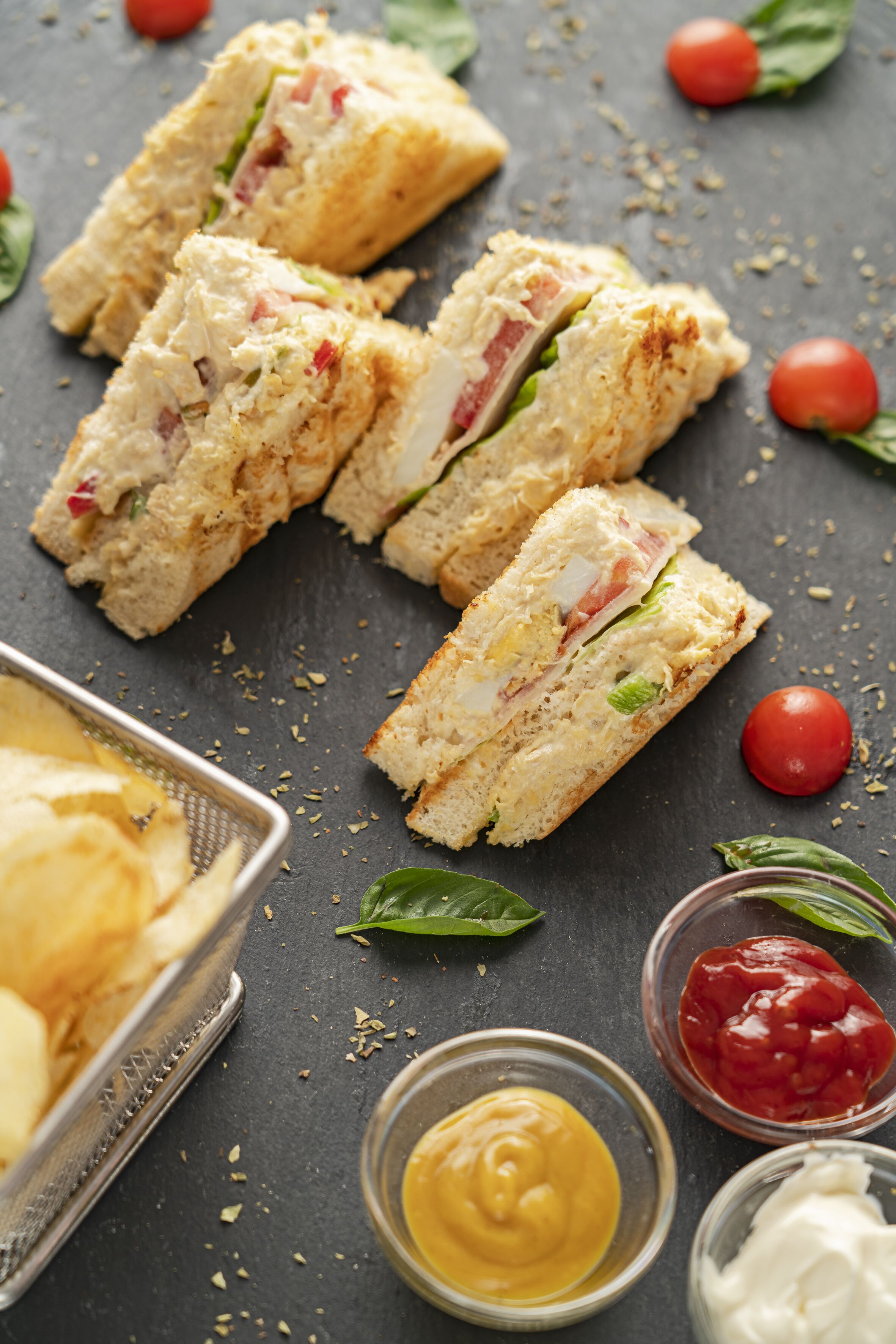 Un sandwich coupé en morceaux, accompagné d’une sélection de condiments et d’un panier de chips.