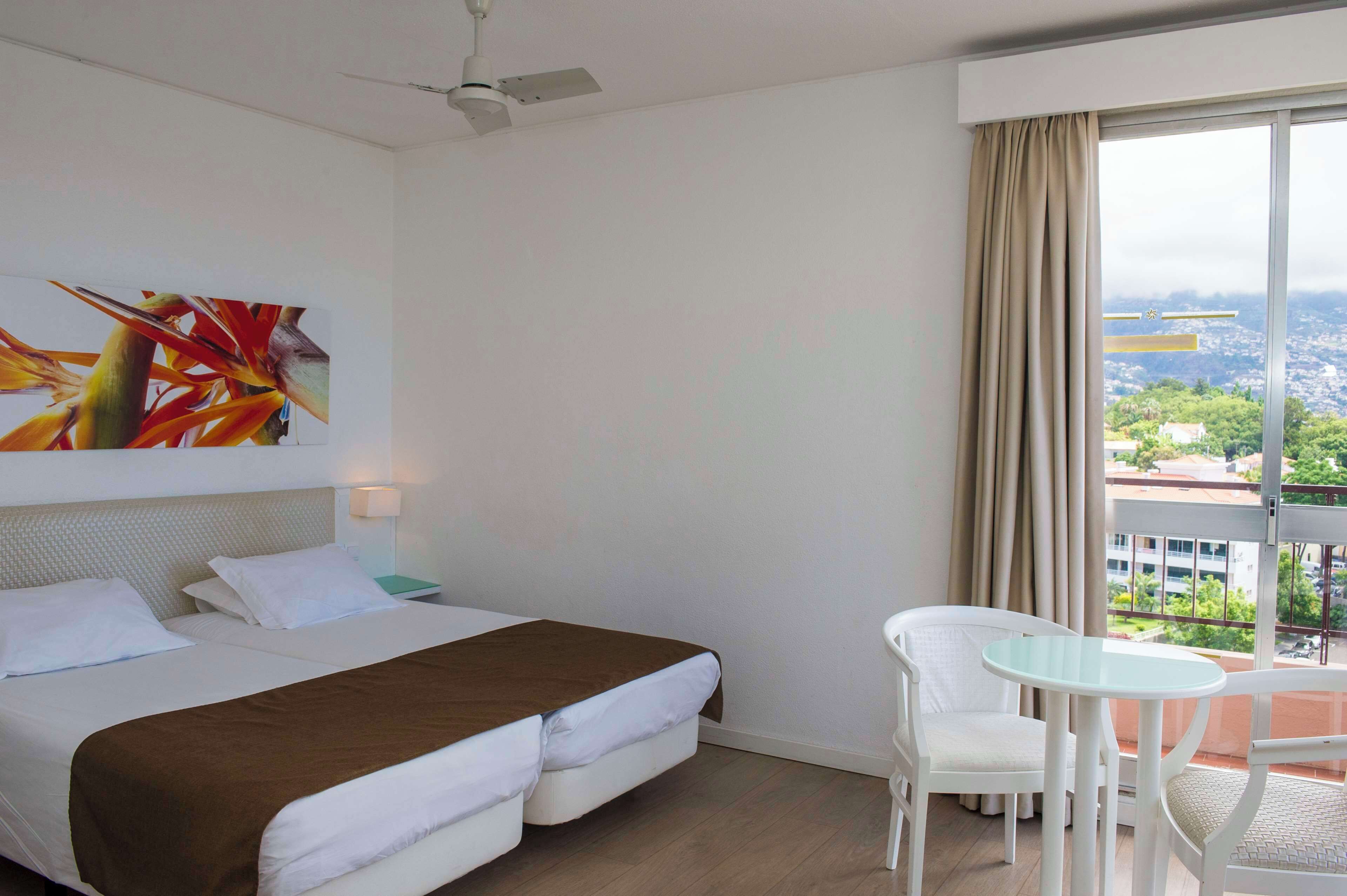 Habitación de hotel con cama, mesita de noche, una mesa con sillas y un ventanal con vistas a la ciudad.