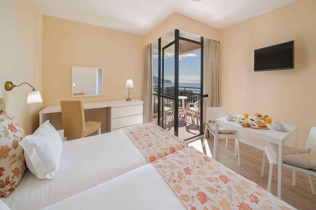 A hotel room with a bed, desk, chair, and balcony with a view.