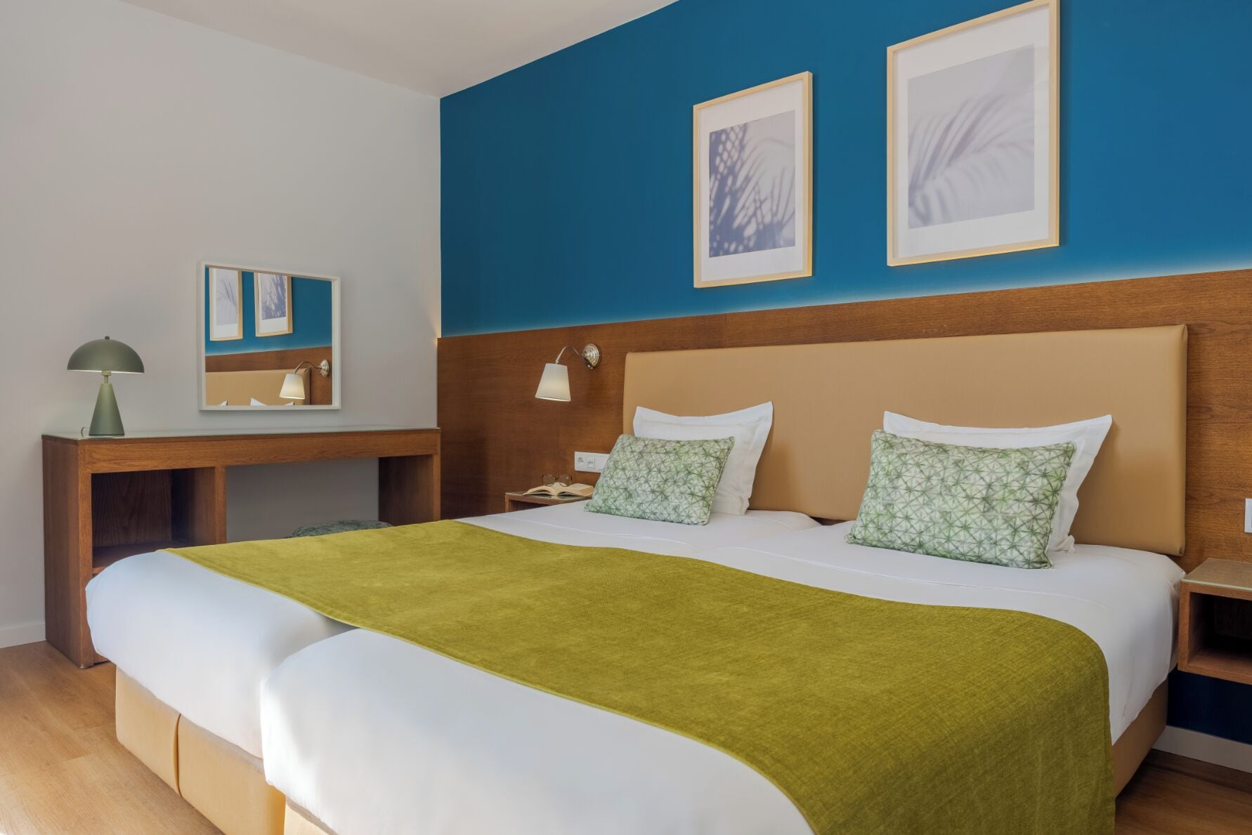 Une chambre d’hôtel avec un grand lit, une table de chevet en bois, un bureau avec une lampe et deux tableaux encadrés au mur.