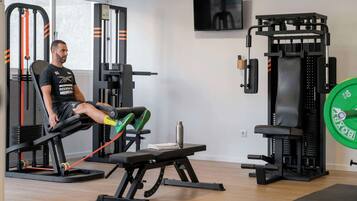 Un gimnasio con una prensa de banca, estantes para pesas y un televisor en la pared.