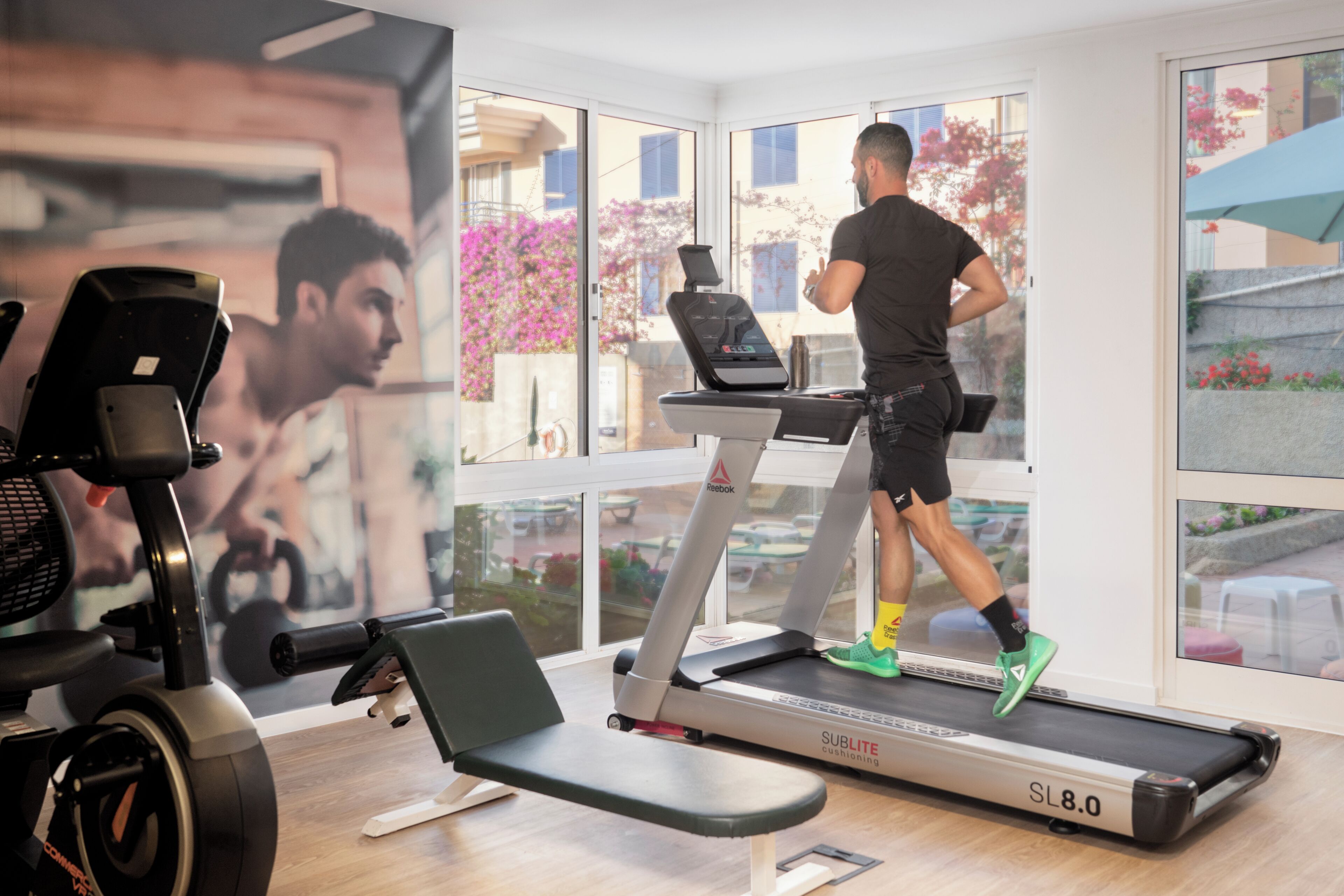 Ein Fitnessstudio mit Trainingsgeräten, darunter Laufbänder und Ergometer, und großen Fenstern mit Blick ins Freie.
