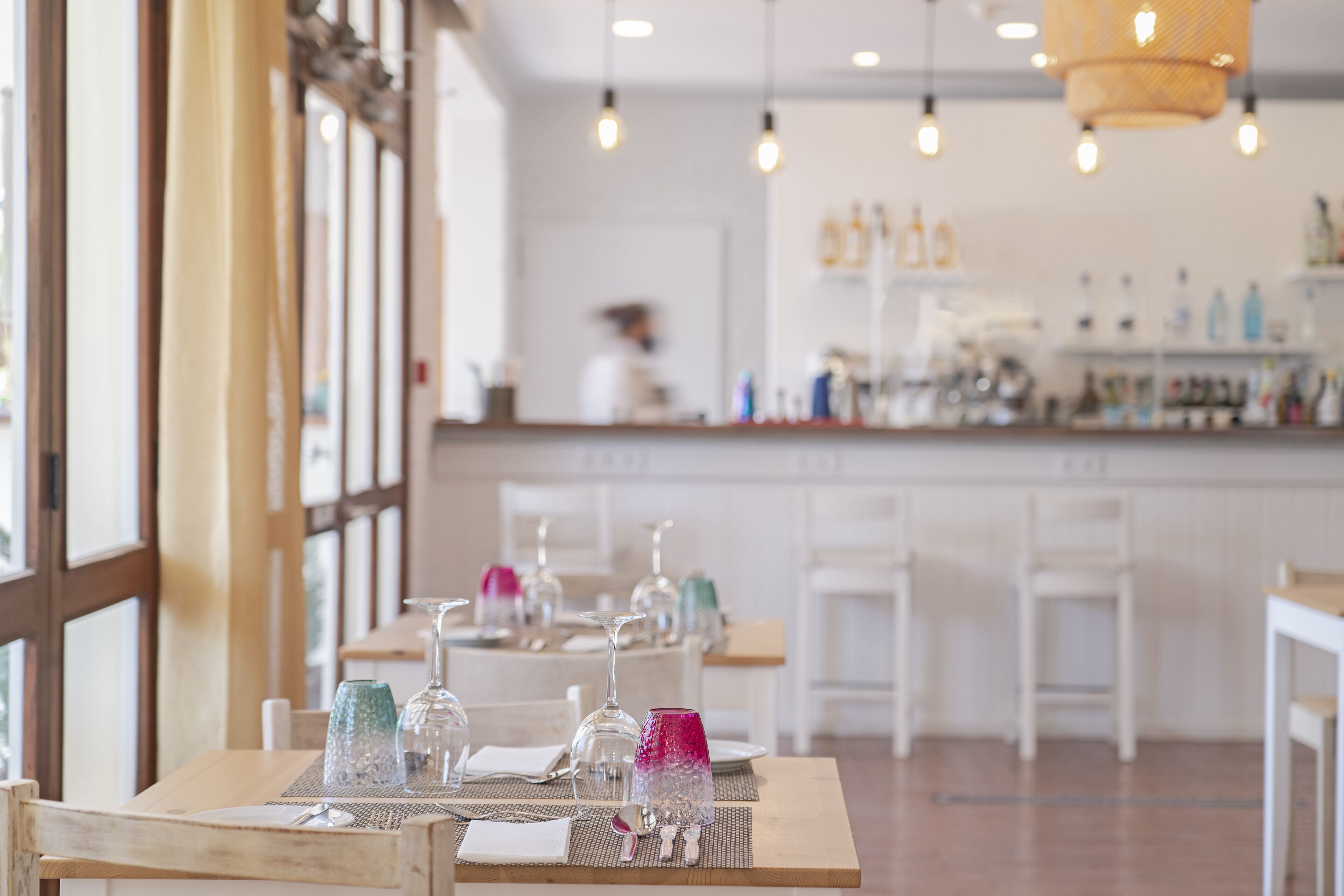 Un ristorante con tavoli apparecchiati per consumare i pasti, un angolo bar sullo sfondo e ampie finestre che lasciano entrare la luce naturale.
