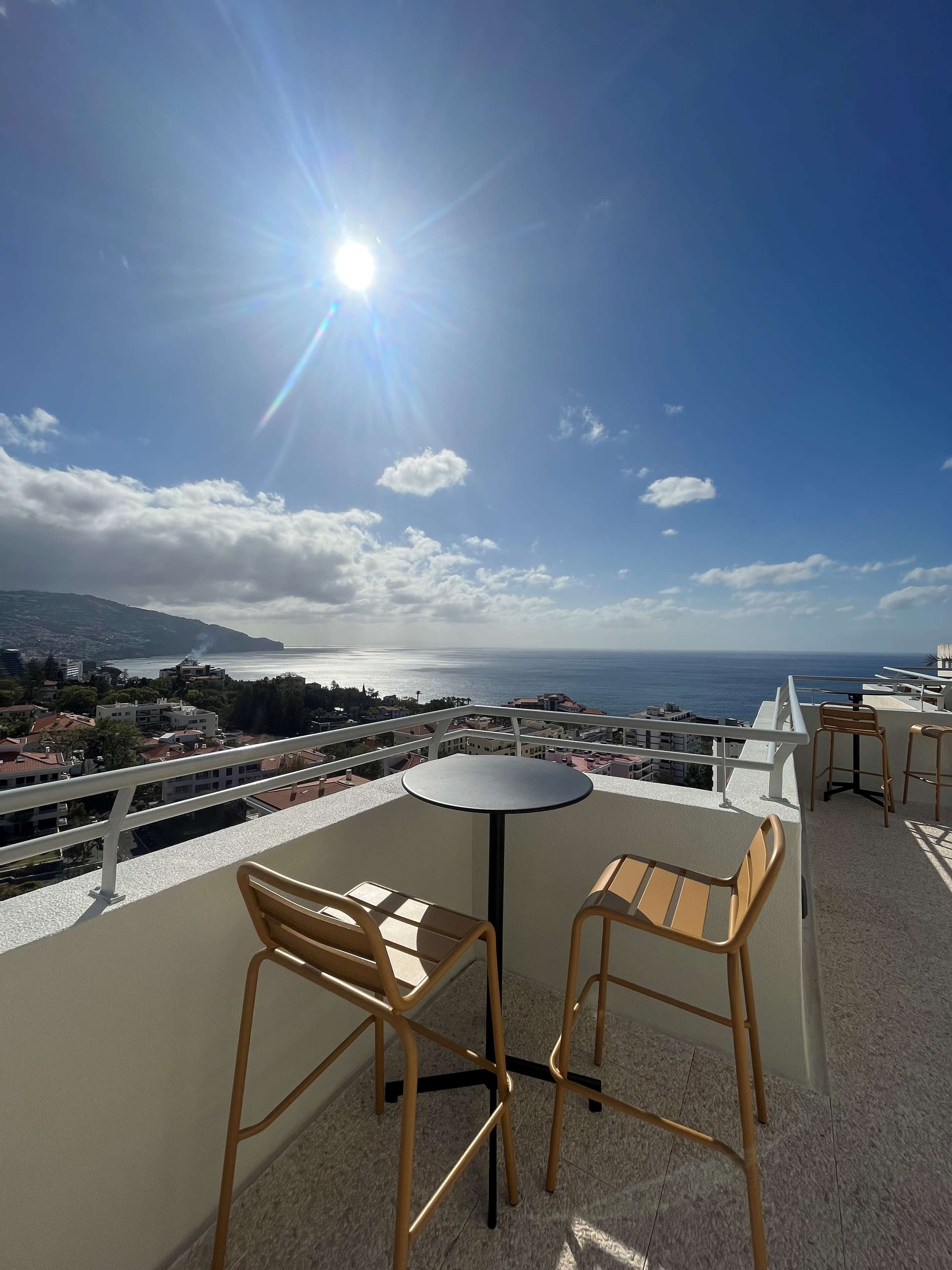 A balcony with a round table and two chairs, overlooking a scenic view of the sea and distant land.