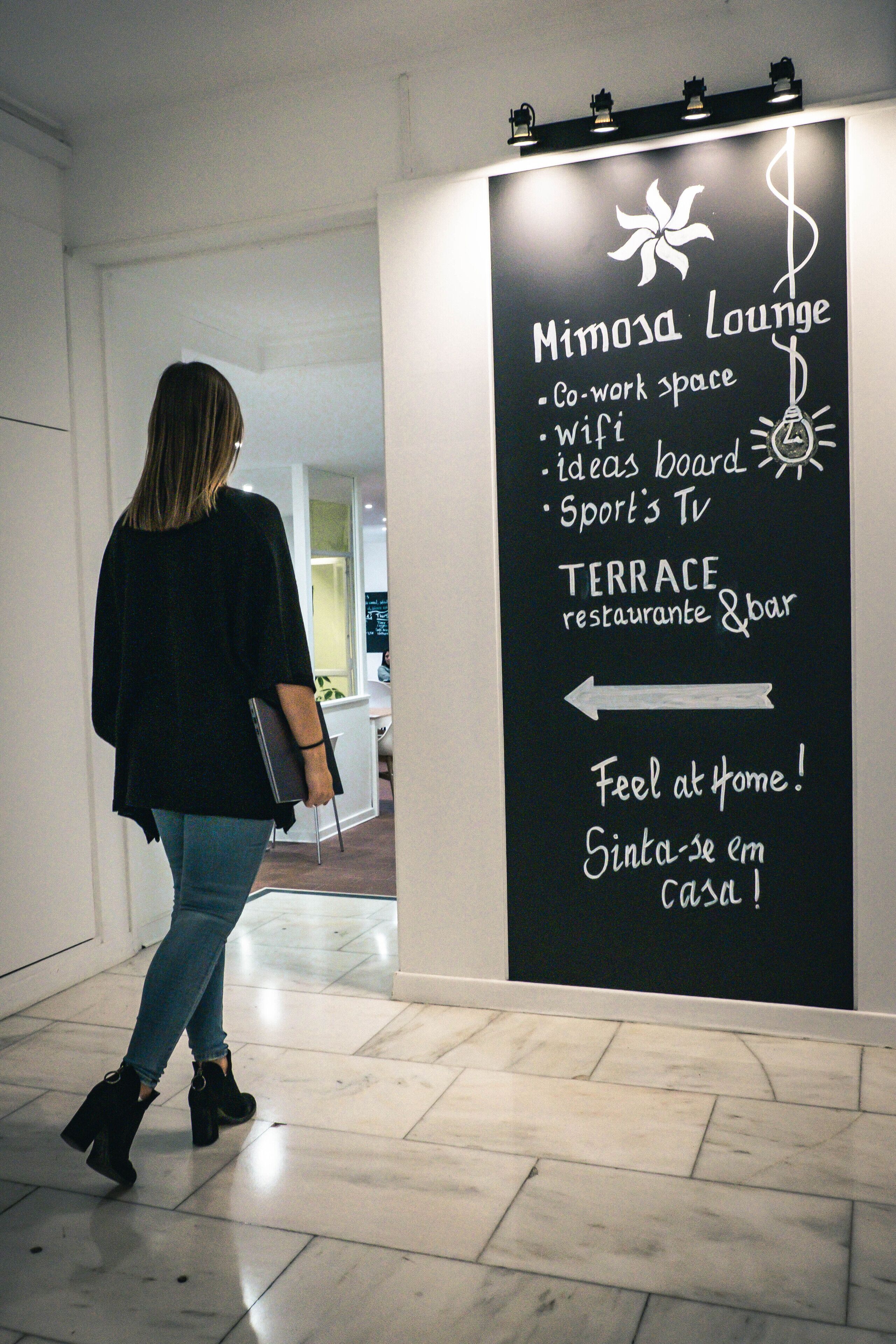 A woman walking towards a signboard with amenities listed such as co-work space, wifi, ideas board, sport's TV, and terrace restaurant & bar.