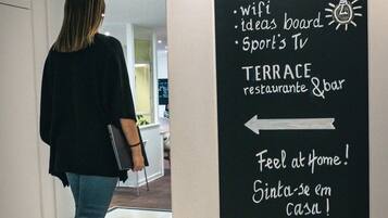 A woman walking towards a signboard with amenities listed such as co-work space, wifi, ideas board, sport's TV, and terrace restaurant & bar.