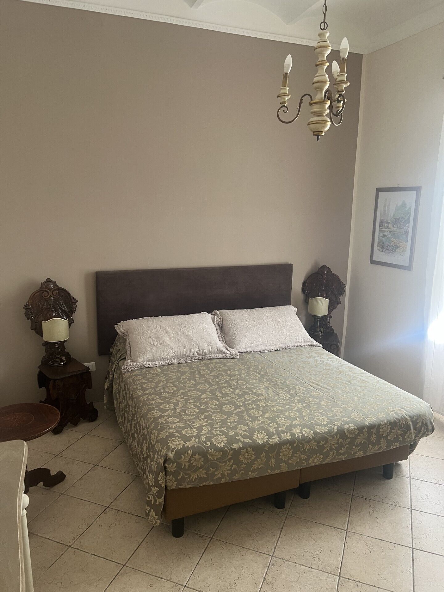 Une chambre à coucher avec un grand lit, deux tables de chevet avec des lampes, un lustre et un tableau encadré au mur.