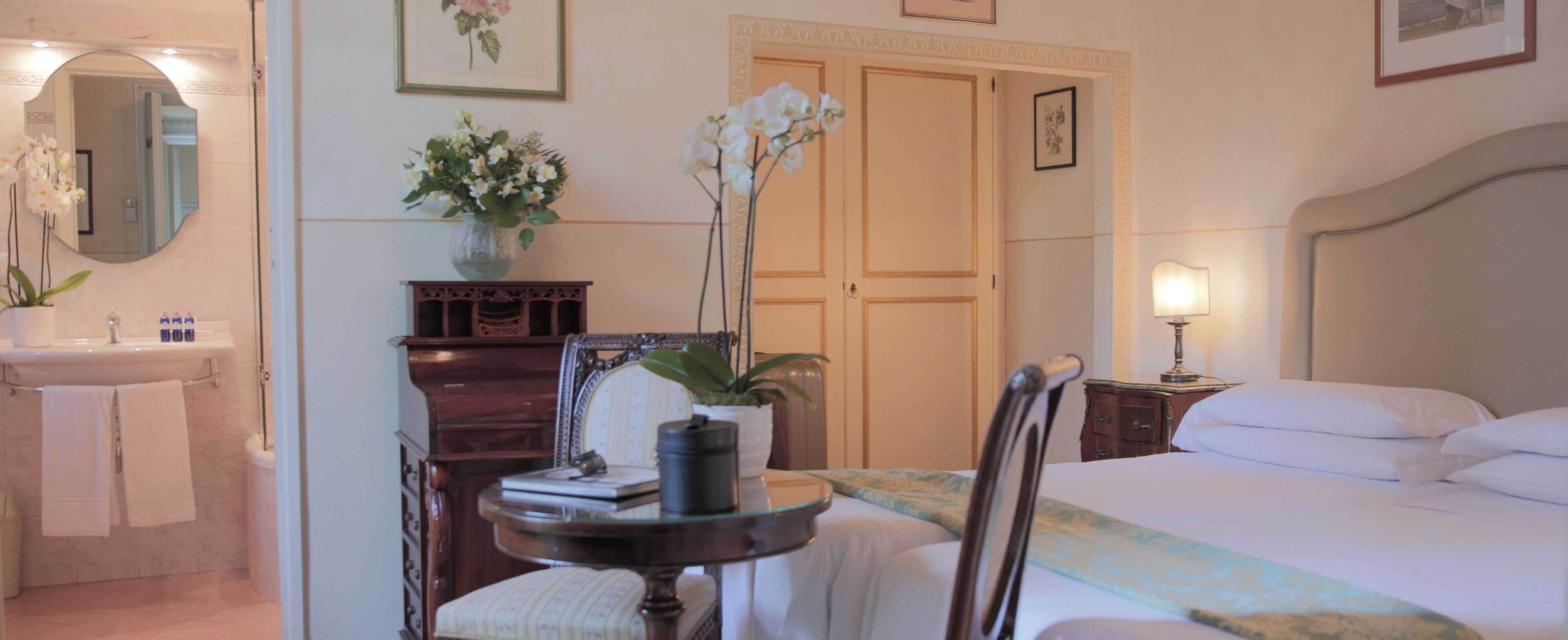 A hotel room with a bed, a bedside table with a lamp, a chair, a desk with a vase of flowers, a dresser, a mirror, and a bathroom with a sink and towels.