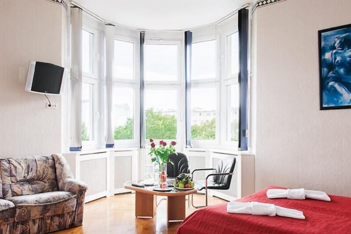 A hotel room with a bed, a desk with a vase of flowers, a chair, a sofa, and large windows.