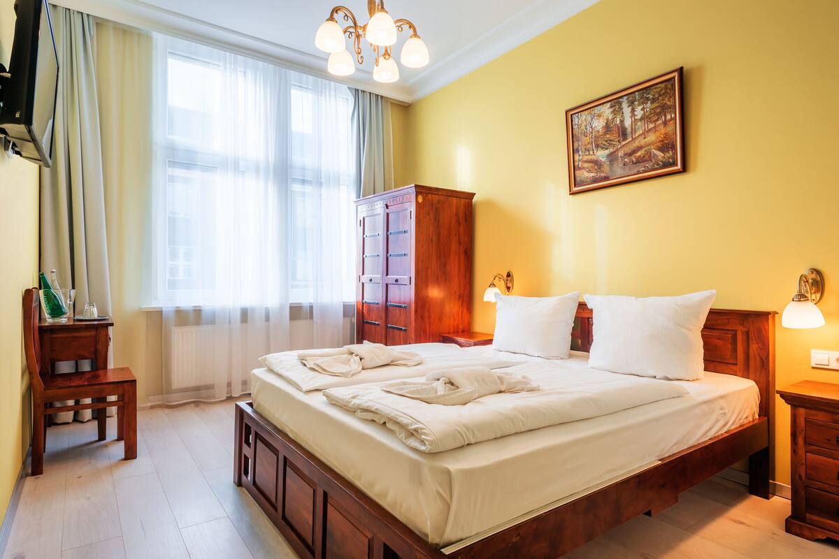 Ein ordentlich bezogenes Bett mit weißen Leinen und Handtüchern, ein Nachttisch aus Holz mit Lampe, ein Holzschrank und ein Bild an der Wand.