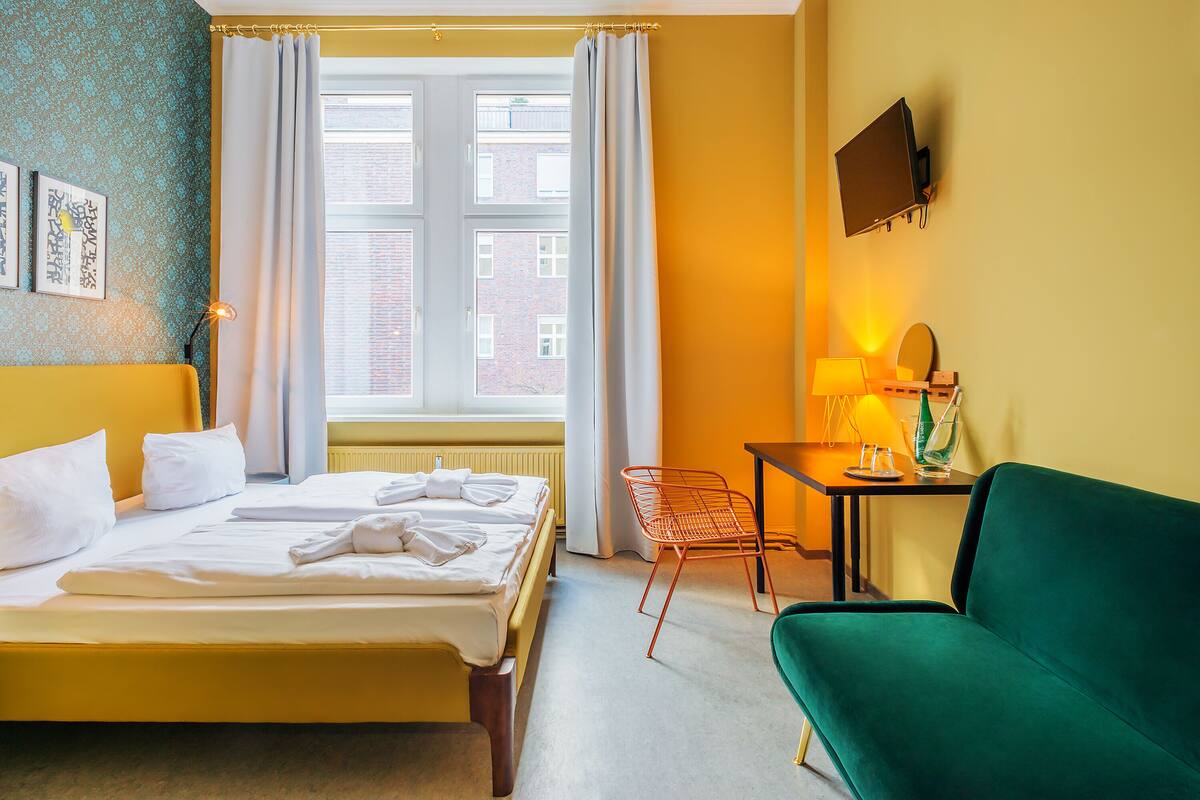 Ein Hotelzimmer mit Bett, Schreibtisch, einem grünen Sofa, einem an der Wand montierten Fernseher und einem Fenster mit Vorhängen.