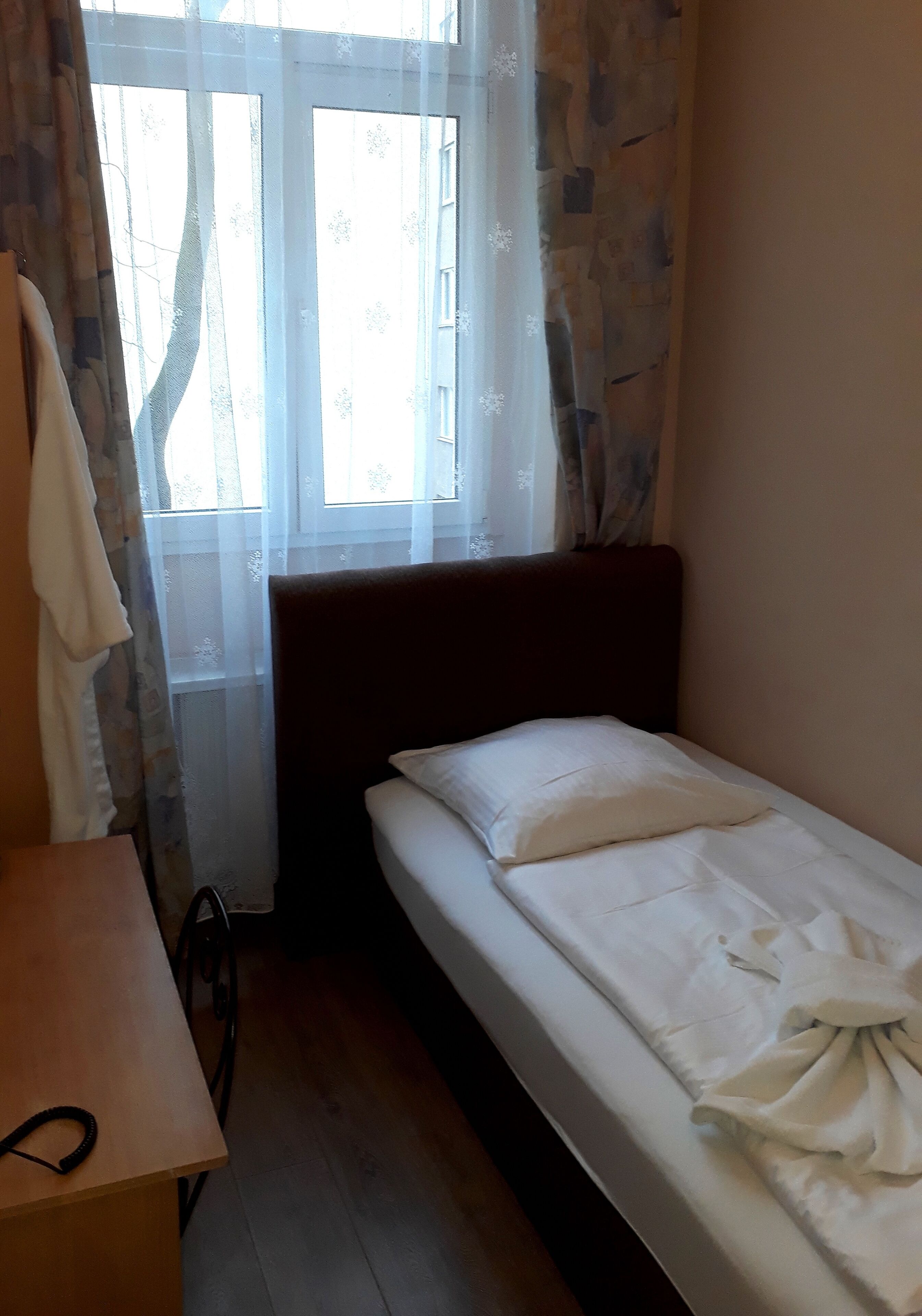 Une petite chambre avec un lit simple, une tête de lit en bois, une table en bois et une fenêtre ornée de rideaux légers.