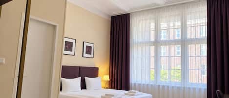 A hotel room with a large bed, a chandelier, a window with curtains, and a door.