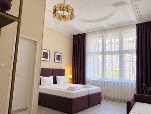 A hotel room with a large bed, a chandelier, a window with curtains, and a door. - Hotel Bellevue am Kurfürstendamm (Berlin)